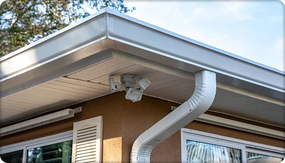 White gutter and curved downspout installed on stucco home with soffit-mounted security camera